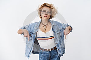 Questioned clueless cute young woman with short blond hair in transparent glasses and denim jacker pointing down with