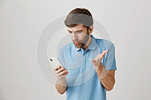 Questioned good-looking guy looking at screen of smartphone, lifting hand cause he is clueless and annoyed with what he