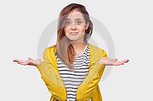 Questioned young woman, looking uncertain during discussing, dressed in striped t-shirt and yellow cardigan, posing over white