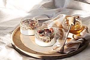 Quick healthy breakfast greek yogurt, honey, granola with dried berries and nuts in glass jar on wooden tray on white bedding