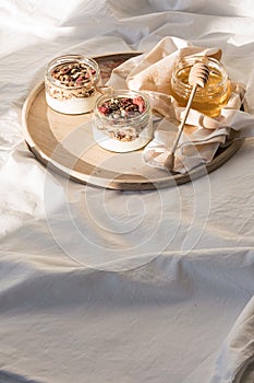 Quick healthy breakfast greek yogurt, honey, granola with dried berries and nuts in glass jar on wooden tray on white bedding