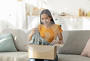 Quick purchases in online store. Satisfied asian woman opening carton box sitting on sofa in living room at home