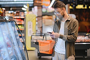 Quick text during shopping. Handsome young man holding mobile phone and smiling while standing in a food store