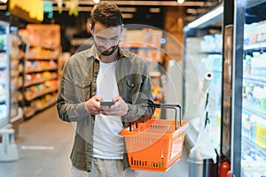 Quick text during shopping. Handsome young man holding mobile phone and smiling while standing in a food store