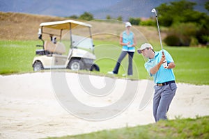 In and quickly out of a bunker. A young male golfer chipping his ball out of a bunker while his female partner looks on