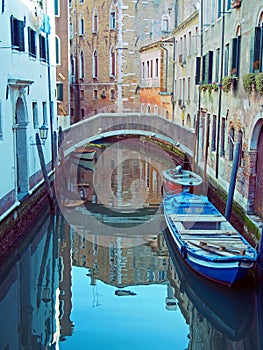 Quiet narrow canal crossed by a bridge with moored boats and old buildings reflected in the water in Venice