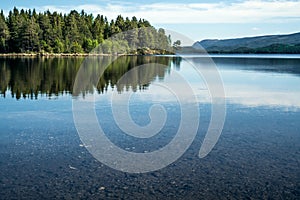 A quiet peaceful summer morning with view over crystal clear calm lake with pebbles in the bottom and forest reflected