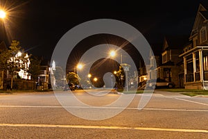 Quiet suburban street at night - illuminated by glowing streetlights - houses lined up - parked cars - clear, starless sky