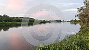 On a quiet summer evening, after sunset, the sky with clouds over the river turns pink and is reflected in the calm water. Shrubs