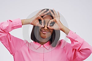 Quirky, funny face and finger glasses with an indian woman in studio on a gray background looking silly or goofy. Comedy