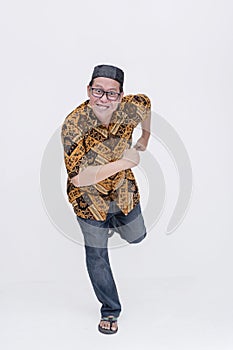 A quirky middle aged man runs towards the camera in an exaggerated and funny way. Wearing a batik shirt and songkok skull cap with