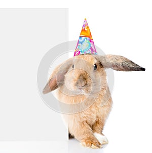 Rabbit in birthday hat peeks out from behind a blank banner. Isolated