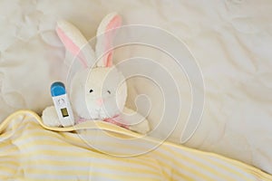 Rabbit with thermometer under paw lies in a crib on a white bed. toy got sick. Playing doctor. Protection from viruses, influenza