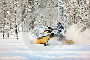 Racer in the outfit of a jumpsuit and a helmet, driving a snowmobile by the deep snow surface on background of snowy forest.