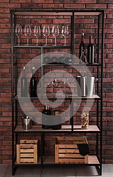Rack with bottles of wine and glasses near brick wall