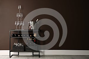 Rack with bottles of wine and glasses near brown wall, space for text