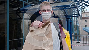 Rack focus from male courier in eyeglasses and coronavirus face mask to stretched package with takeaway food. Portrait