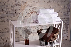 Rack with a stack of three white color towels and baskets with clean brown towels and toilet decor near a brick wall. Shelf with