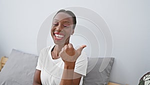 Radiant african american woman relaxing on her comfy bed, confidently pointing to the side with a thumbs-up, expressing cheeriness