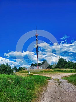 A radio mast on a hill surrounded by trees and a meadow. Cell phone tower and remote station receiver transmitter.