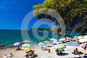 Resting people on public finely pebbly and sandy beach at a sunny summer day. Rafailovici, Montenegro