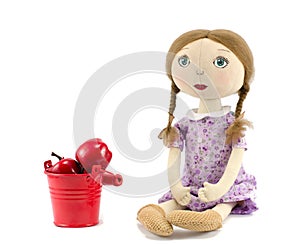 Rag-doll girl with blond hair dressed in purple and bucket with red apples on white background