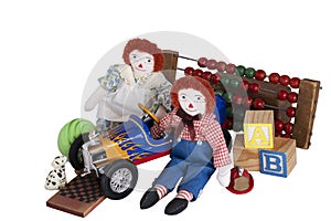 Raggedy Ann & Raggedy Andy sit with old toys: race car, checkers, blocks, counter, ball, fire hat & dog cutout.