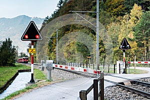 Railraod crossing with blinking warning lights and a red train approaching in the Swiss countryside