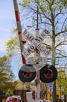 A railroad crossing signal with tall white and red arms surrounded by lush green trees an parked cars in the Marietta Square