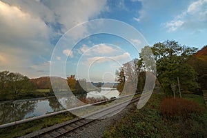 Railroad tracks on a river with a forest in autumn under a blue sky with clouds