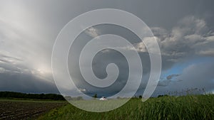 Rain clouds are a strip of deciduous forest and green grass at the edge of a field and a white car, panoramic landscape