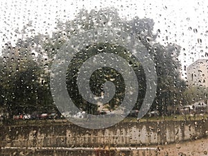 Rain drops on wet window and street tree, blue sky and stormy rain clouds behind, blurred city bokeh. Concept of rainy