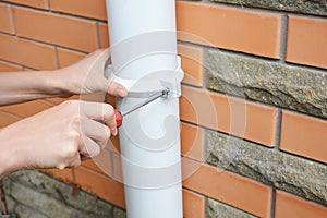 Rain gutter installation: a man is screwing the downpipe bracket, socket clip with a screwdriver to fix the downspout to the wall