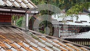 Rain water is falling and flows down the tiled roof into the drainpipe. A green yard with pine trees in the background.
