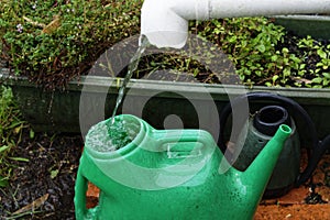 A green plastic watering can is being filled from a rain water spout, it is collecting the rain water