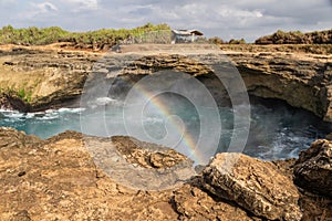 Rainbow above water at Devil`s Tear, Nusa Lembongan. Rocks in foreground. Cloudy sky.