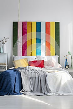 Rainbow bedhead behind white bed with colorful pillows and blankets in bright bedroom interior. Real photo