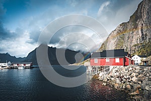 Rainbow ofer red houses rorbuer of Reine in Lofoten, Norway with red rorbu houses, clouds, rainy blue sky and sunny