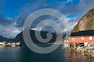 Rainbow ofer red houses rorbuer of Reine in Lofoten, Norway with red rorbu houses, clouds, rainy blue sky and sunny