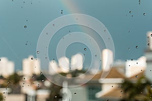 A rainbow over some buildings behind a glass windows with water drops