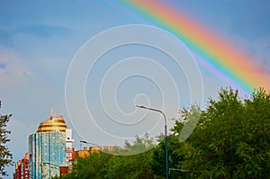 Rainbow after the rain on the blue sky. Foliage of trees. Street lights and building