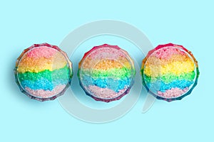 Rainbow shaving ice in glass dessert bowls on light blue background, flat lay