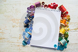 Rainbow spectrum buttons and reels with colorful threads, laid around a notebook with space for text on a light wooden background