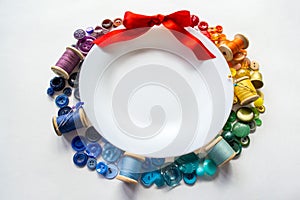 Rainbow spectrum buttons and reels with colorful threads, laid out in form of wreath with red bow around a white plate with space