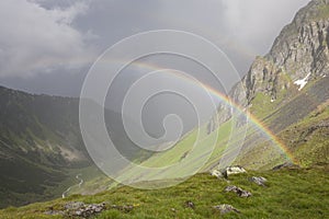 Rainbow stretching over a mountain valley in front of dark thunder storm.