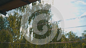 Raindrops flow down in thin streams from the roof of the building on a blurred background of trees and sky