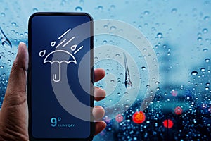 Rainy Day Concept. Hand Holding Smartphone with Weather Information show on Screen. Blurred Traffic Jam and Rain Drops on Glass W