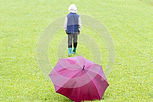 Rainy weather. Little boy go away. Red umbrella on the green grass.