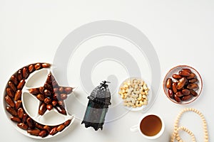 Ramadan dates fruit in crescent moon plate, arabic lantern, nuts, cup of tea, rosary on white table. Ramadan Iftar food. Top view