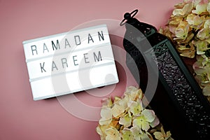 Ramadan Kareem word in light box with black metal lantern and flower decoration on pink background